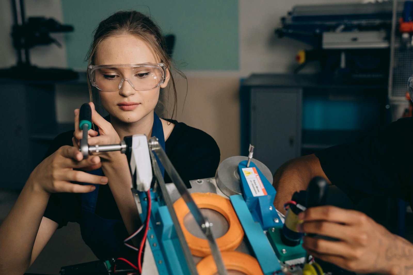 A young woman with safety glasses assembling a robotic device indoors.